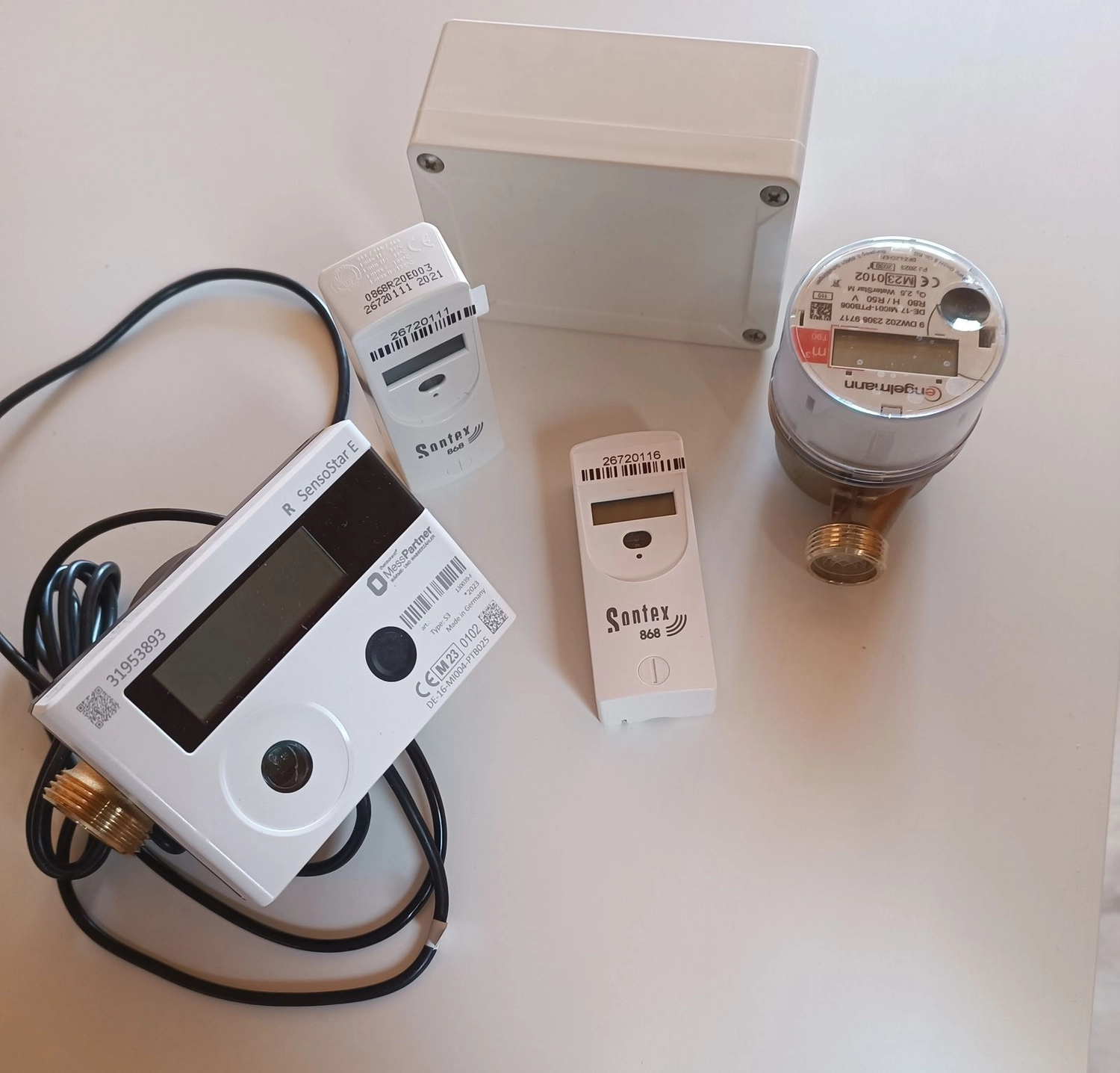 Fair Therm GmbH - Rechtskonforme Abrechnung und Analyse - Ein weißes und ein silbernes Wärmemessgerät mit angeschlossenen Kabeln stehen auf einem Tisch neben zwei kleineren weißen Fernsensoreinheiten.