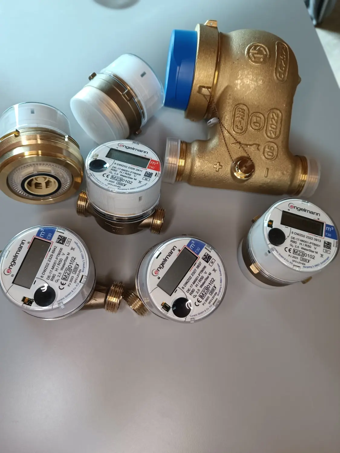 Fair Therm GmbH - Rechtskonforme Abrechnung und Analyse - Eine Sammlung von Wasserzählerkomponenten, darunter Messingarmaturen und mehrere kleine, runde elektronische Wasserzähler mit Digitalanzeigen, einige davon mit der Aufschrift "Cengelmann".